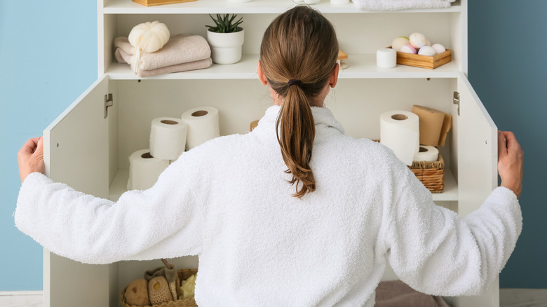 Woman looking in bathroom cabinet filled with toilet paper, towels, and toiletries