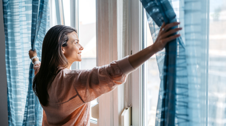 A smiling woman is pulling back blue curtains from a window