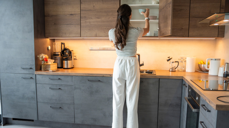 Woman standing in a kitchen reaching into an upper cabinet
