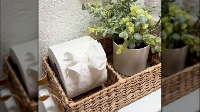 Toilet paper embellished with bow sitting in stylish basket alongside a potted fake plant