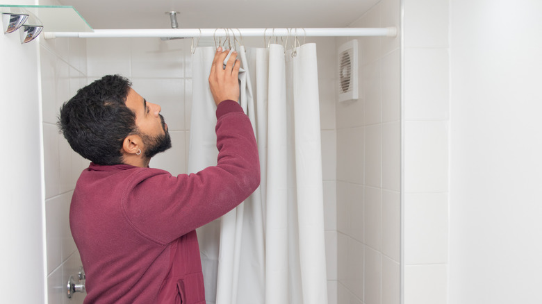 A bearded man wearing a maroon fleece jacket stands in a white tiled bathroom, installing or adjusting a white shower curtain on the shower rod
