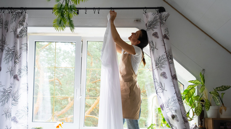 Woman in apron removes curtains, big window with plants in background
