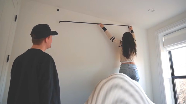 Woman hanging a curtain rod behind a bed on empty bedroom wall for DIY headboard