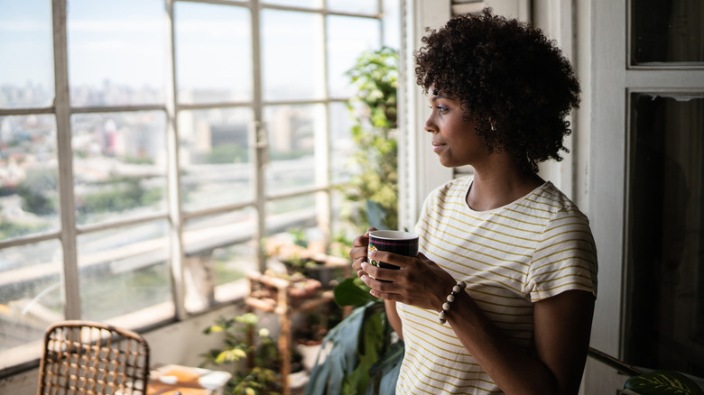 A woman holds a coffee mug while looking out a large window