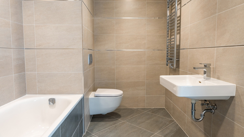 beige and grey tiled bathroom with a mounted white sink, toilet, and tub
