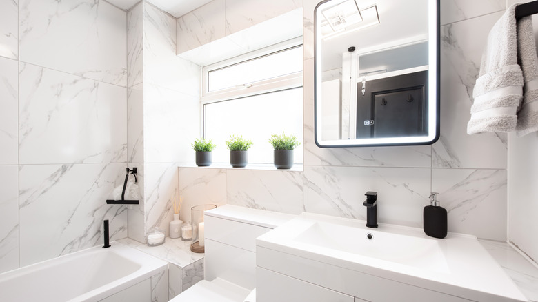 mostly white small bathroom with marble tiles, bathtub, sink, and toilet as well as window with plants in it
