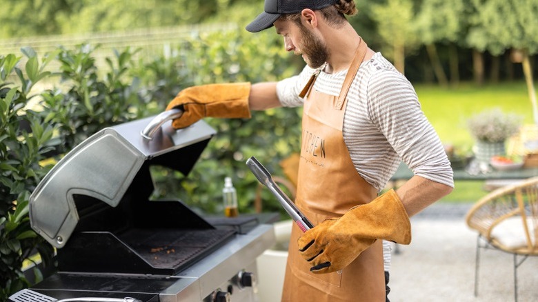 Man in apron and gloves holding tongs while opening propane grill on patio