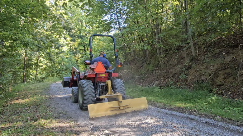 Person using scraper blade tractor attachment on gravel driveway.