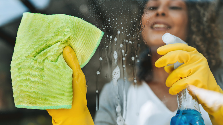 Woman sprays Windex on a glass window while holing cleaning cloth in hand