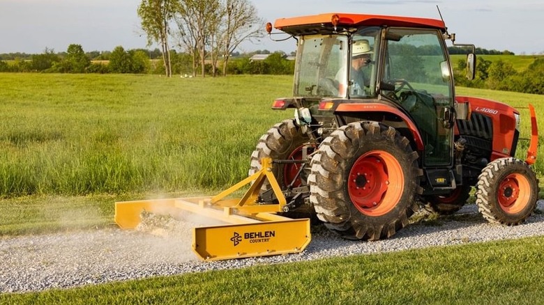 A person driving an orange tractor pulling a grader or land plane behind it to smooth a gravel drive.