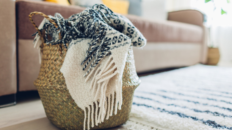 A throw blanket in a woven basket on the floor with a rug next to a couch