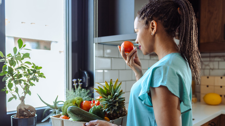 A woman holds a box of vegetables and smells a tomato