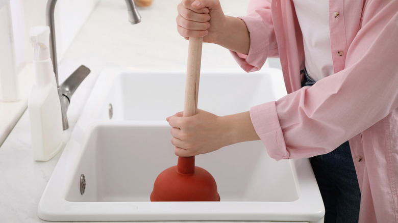 Person wearing a pink shirt and holding a plunger in a white sink
