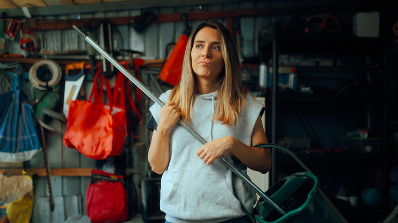 A skeptical-looking person holds a mop and bucket in a messy garage