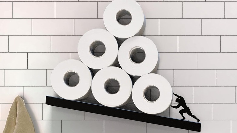 Black floating wall shelf with Sisyphus figure on lower end holding up six rolls of toilet paper