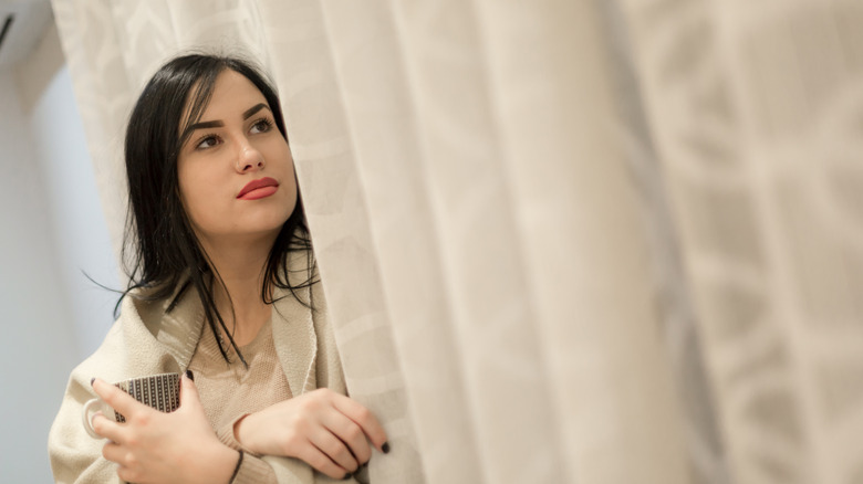 A woman sits by curtains, holding a mug and thinking