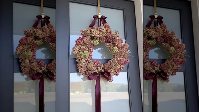 A pink and green hydrangea wreath with burgundy bows on a blue/grey front door