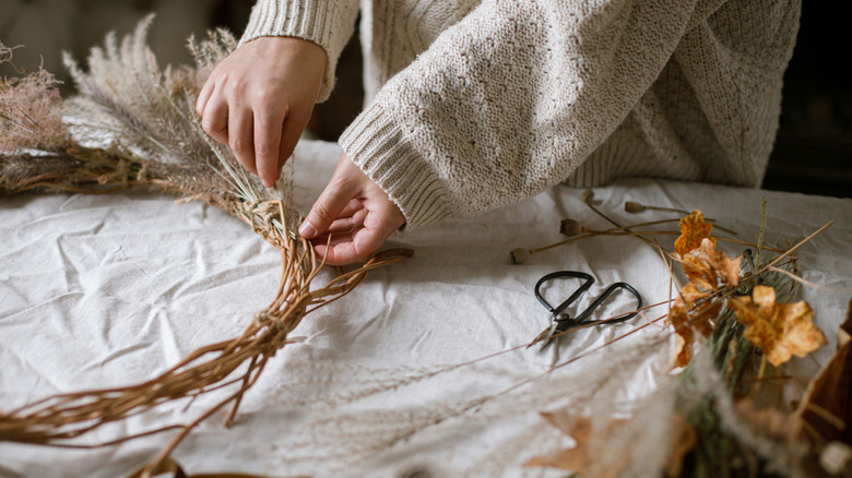 A person in a beige sweater tucking floral picks into a grapevine wreath to decorate for fall.