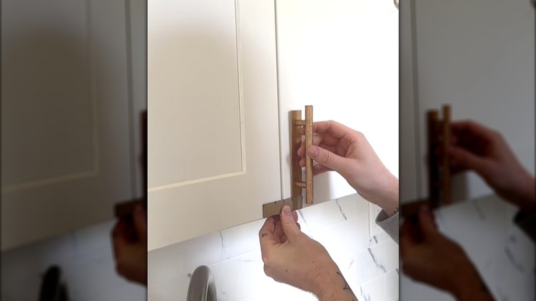 Hands attaching gold cylindrical handles to white modern cabinets in kitchen