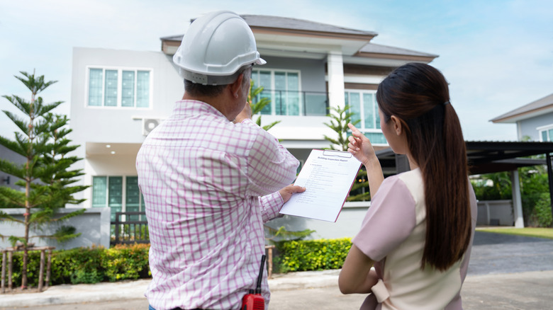 A building inspector wearing a hardhat and holding a clipboard stands next to a homeowner while looking at the exterior of a house