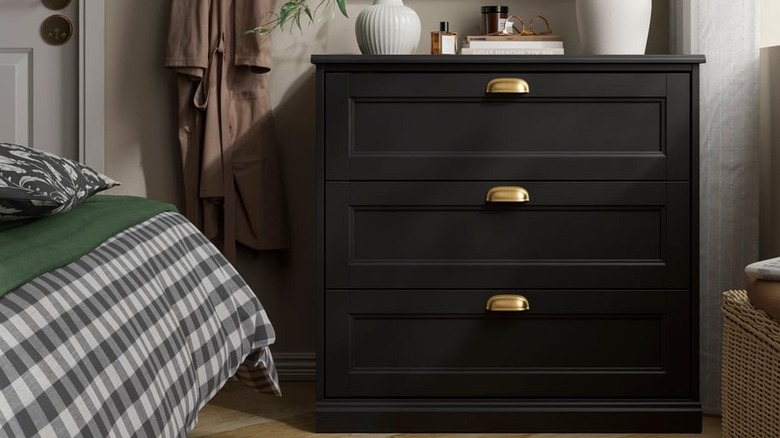 A black three-drawer dresser sits near a plaid bedspread