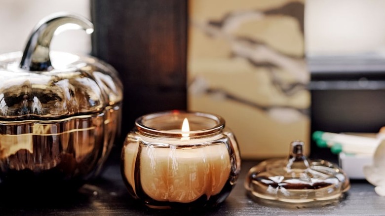 A lit candle in a pumpkin-shaped glass jar sits on a wooden ledge