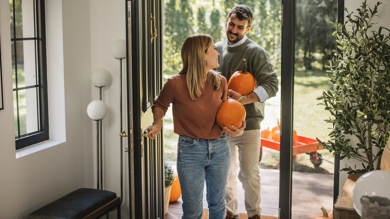 A young couple walks through the front door of a minimalist home carrying pumpkins