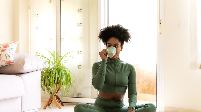 A woman drinks from a mug while sitting in front of a spider plant.
