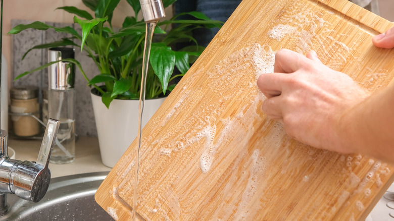 Hands washing a bamboo cutting board with soap and water.