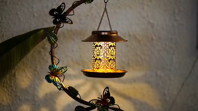 A solar light glowing on the middle tier of a bird feeder, hanging from a metal chain and framed by a decorative screen.