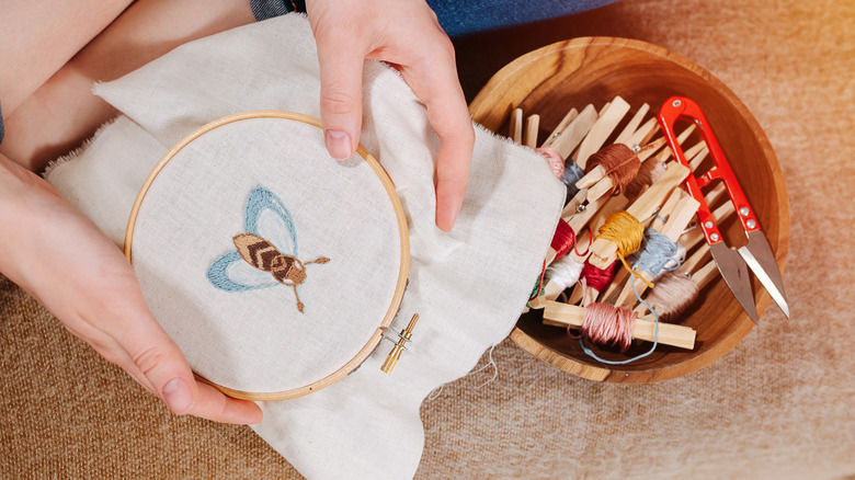 woman holding a sampler with an embroidered bee and embroidery thread in a bowl on the floor