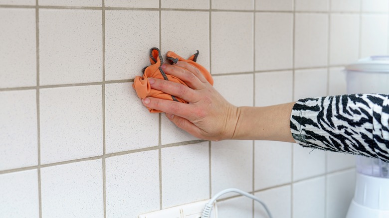 A person wiping down white tiled backsplash with orange cloth