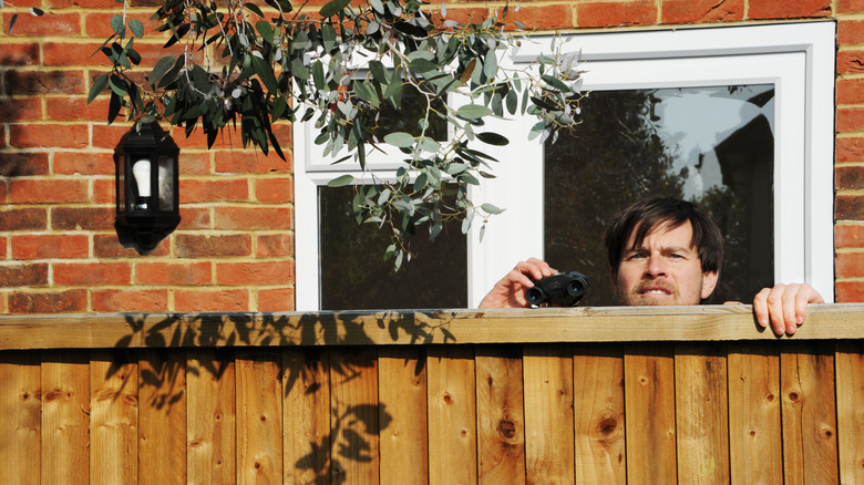 A creepy neighbor with binoculars peers over a wooden fence