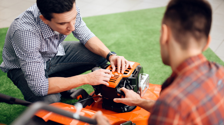 Two men look at an orange lawn mower together on a strip of grass, inspecting it