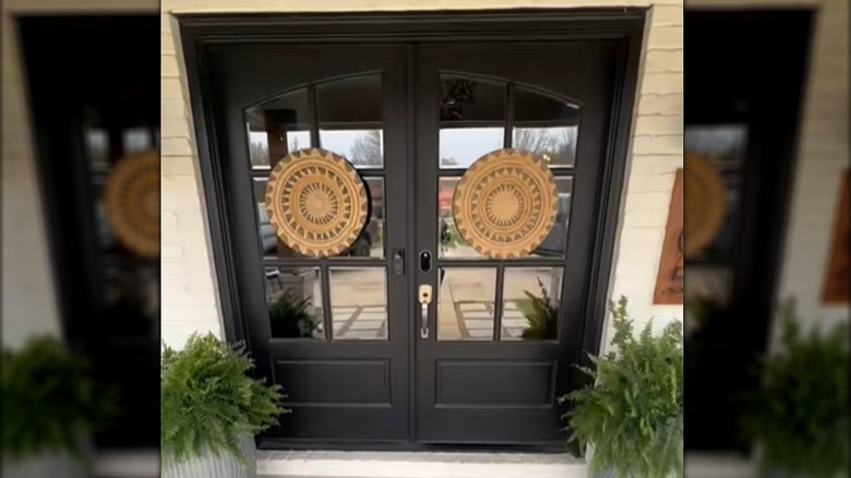 Decorative woven disks positioned over the windows on black double front doors with plants next to the entrance