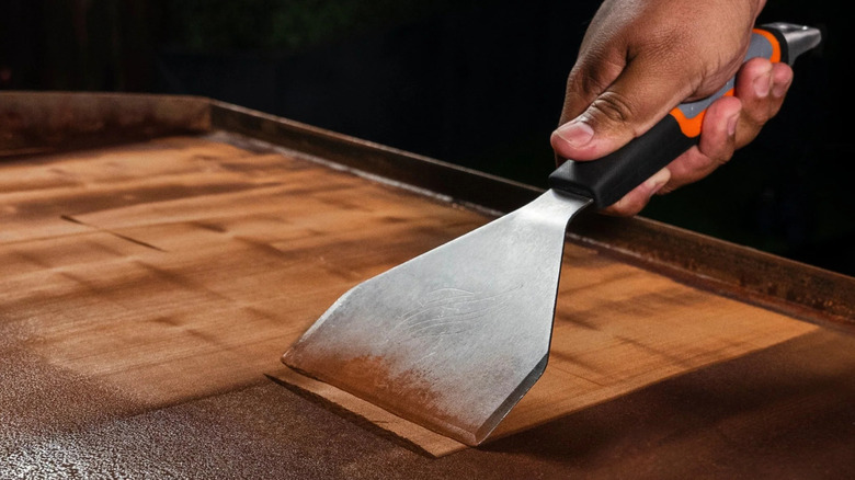A person uses a scraper to remove rust from a Blackstone griddle