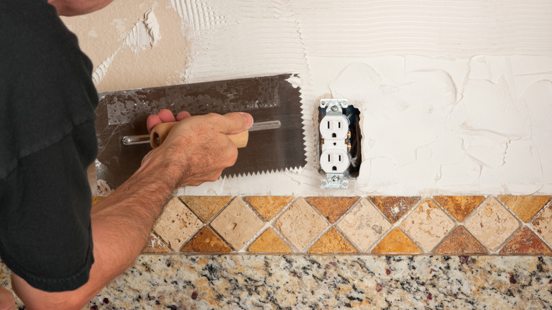 A person is applying a Tuscan diagonal style backsplash in earth tones against a wall.