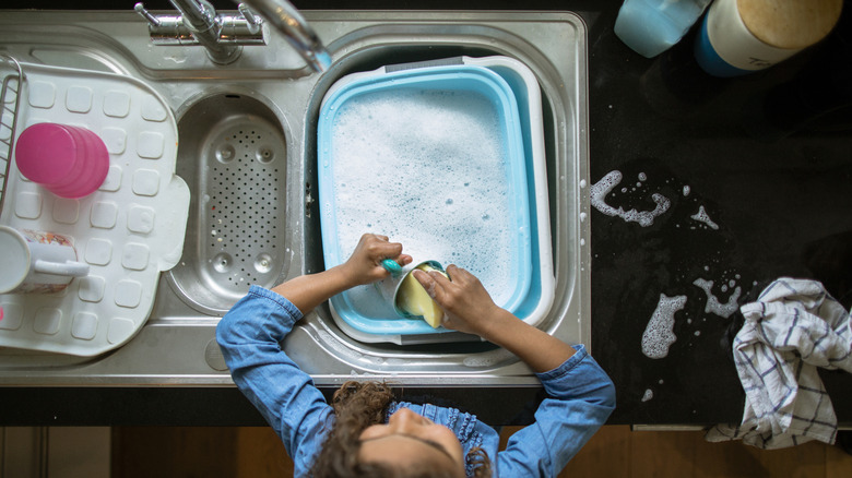 A young girl washing dishes in a plastic basin in the kitchen sink
