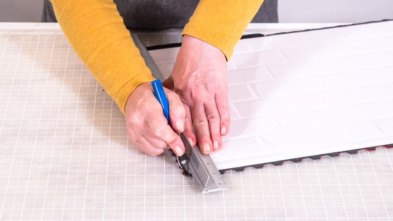 A person uses a utility knife and a straight-edge to cut white peel-and-stick tile