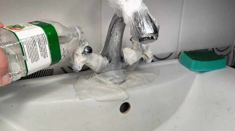 A faucet tap is covered in paper towels with a hand pouring vinegar on top
