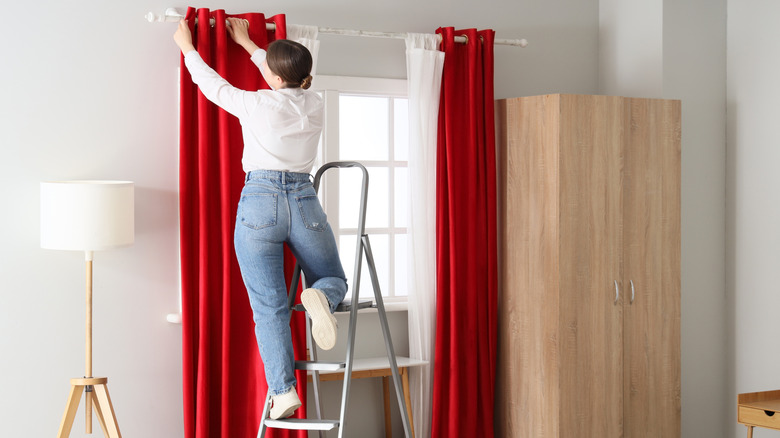 A woman stands on a ladder and adjusts red curtains hanging above a bedroom window