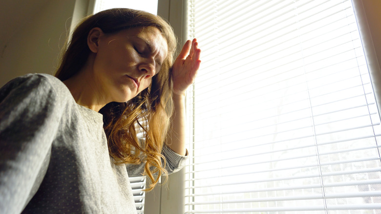 A woman shielding her eyes in a sunny window with blinds