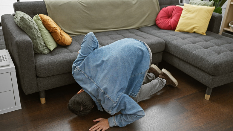 Young man in jean jacket looking for lost item under gray mid-century modern couch with colorful pillows