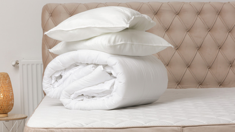 White folded comforter and two pillows stacked on a bed with beige headboard