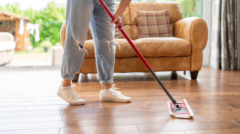 A person mopping a hardwood floor with a brown couch in the background