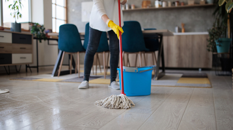 A woman wearing yellow gloves mopping a kitchen floor with a bucket behind her
