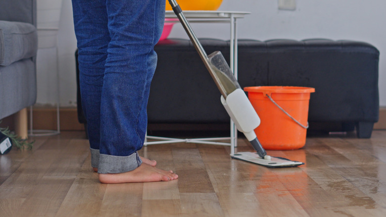 A person mops laminate flooring next to furniture and an orange bucket