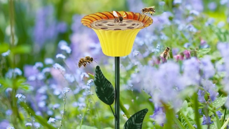 Bees hover above a yellow flower-shaped birdbath surrounded by purple flowers