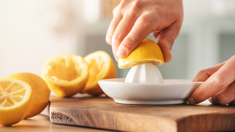 A hand squeezing half of a lemon on a white juicer placed on a wooden cutting board