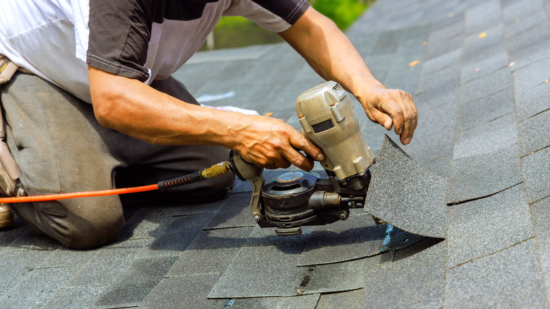 A kneeling handyman uses a pneumatic gun to nail asphalt shingles to a roof